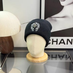 May include: A black ribbed knit beanie featuring a silver-toned metallic cross emblem centered on the front. The emblem is surrounded by a circular border with the text CHROME HEARTS engraved in a gothic-style font.