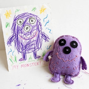 May include: A purple felt monster plush toy with three black button eyes and a stitched pink mouth, displayed next to a white paper drawing of the same purple monster with the text MY MONSTER! :) written in pink crayon at the bottom.