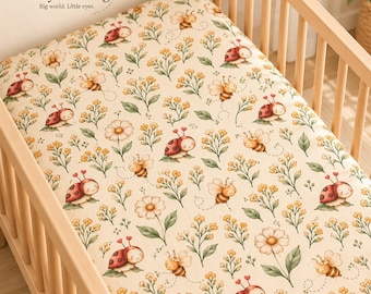 May include: A cream-colored crib sheet featuring a repeating pattern of red ladybugs, yellow bees, and small white and yellow flowers with green stems. The text BabyLullaBug Big world. Little eyes. is printed in the top left corner.