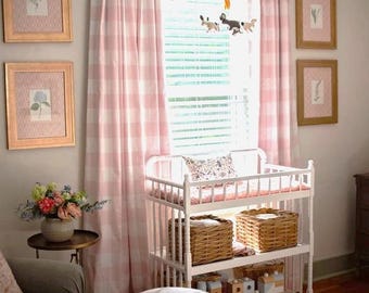 May include: White changing table with two shelves and a pink and white gingham curtain in the background. The changing table has a white changing pad and two wicker baskets on the bottom shelf. There are wooden toys on the top shelf.