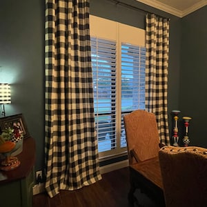 May include: A window with white plantation shutters is flanked by black and white checkered curtains. The room has dark blue walls and hardwood floors. Two chairs with brown upholstery are positioned in front of a wooden table.