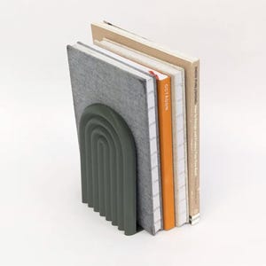 Op de afbeelding: Een donker olijfgroene metalen boekensteun in een geribbelde boogvorm die vier boeken ondersteunt. De boeken hebben een grijze textuurkaft, een dunne oranje rug met het label OCTAGON, een witte rug met rasterpatroon en een beige rug met tekst.