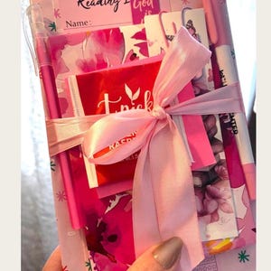 May include: A pink stationery set tied with a light pink satin ribbon, featuring a notebook with a floral pattern, a bookmark labeled Reading God is, a tea bag, and two pink pens. The items are arranged together as a bundled gift package.