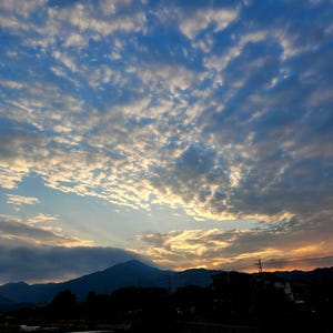 May include: A sunset sky filled with scattered white and gray clouds over a dark mountain silhouette. The horizon glows with orange and yellow light, contrasting with the deep blue upper atmosphere and the shadowed landscape in the foreground.