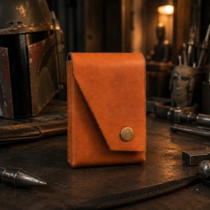 May include: A rectangular, orange leather wallet with a flap closure and a brass button. The wallet is a solid color and appears to be handmade. The background includes a helmet and various tools.
