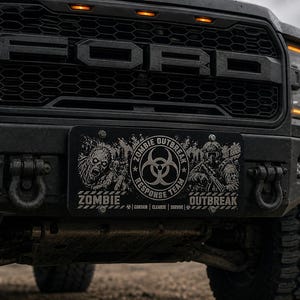 May include: Close-up of a black Ford truck front end. The grill has the Ford logo. A license plate reads "ZOMBIE OUTBREAK RESPONSE TEAM" with zombie and soldier graphics. The truck has a black bumper and large tires.