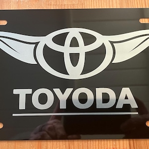以下が含まれることがあります： 黒色のナンバープレート。白い文字で「TOYODA」と書かれ、その下に翼のような拡張部を持つ銀色のロゴがあります。プレートには4つの取り付け穴があり、反射面があります。