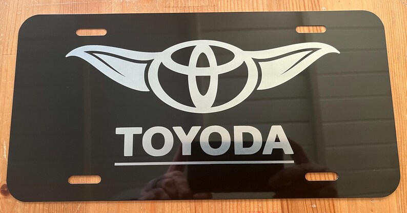 以下が含まれることがあります： 黒色のナンバープレート。白い文字で「TOYODA」と書かれ、その下に翼のような拡張部を持つ銀色のロゴがあります。プレートには4つの取り付け穴があり、反射面があります。