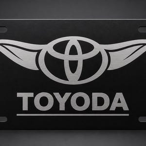 以下が含まれることがあります： 黒色のナンバープレート。トヨタのロゴと「TOYODA」の文字が銀色で表示されています。ロゴは翼のある様式化されたエンブレムです。プレートには4つの取り付け穴があります。