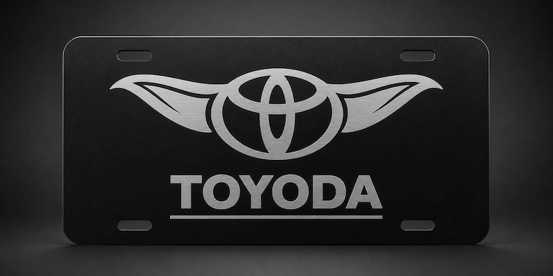 以下が含まれることがあります： 黒色のナンバープレート。トヨタのロゴと「TOYODA」の文字が銀色で表示されています。ロゴは翼のある様式化されたエンブレムです。プレートには4つの取り付け穴があります。
