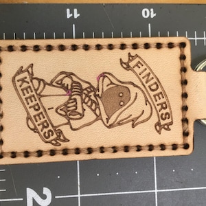 May include: A rectangular, light brown leather keychain with a metal ring. The keychain features a detailed, burnt-in illustration of a hooded figure with the words "FINDERS" and "KEEPERS" on banners.