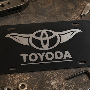 以下が含まれることがあります： 黒色のナンバープレート。トヨタのロゴと「TOYODA」の文字が銀色で表示されています。プレートには4つの取り付け穴があり、工具やハードウェアに囲まれています。