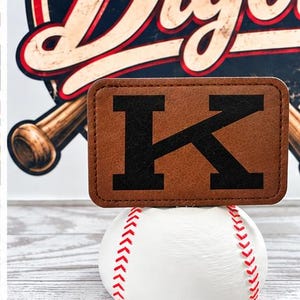 May include: A rectangular brown faux leather patch featuring a large black letter K, resting on top of a white baseball with red stitching. The background displays a graphic with a baseball bat and the partial text Dig.
