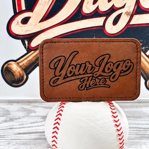 May include: A rectangular brown leather patch with the black text Your logo Here engraved on the surface. The patch is positioned on top of a white baseball with red stitching, set against a background featuring a baseball bat graphic.
