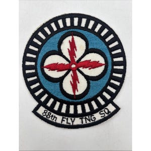 May include: A circular embroidered patch featuring a blue center with a white four-leaf clover shape overlaid by a red lightning bolt design. The border consists of black and white stripes, and a tab at the bottom reads 88th FLY TNG SQ.