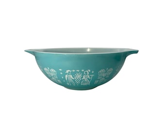 Op de afbeelding: Een turquoise mengkom met twee handgrepen aan de zijkanten, voorzien van een wit gedrukt patroon van figuren, ananassen en gestileerde bloemelementen rondom de buitenkant. De kom heeft een gladde, ronde vorm en een brede, open bovenrand.
