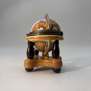 May include: A decorative ceramic globe trinket box. The globe features a map design with brown and white continents. It sits on a tan stand with dark brown legs and a gold-colored handle. The box is hinged and appears to open.