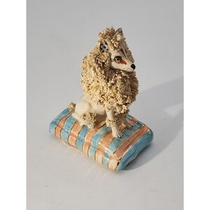 May include: A ceramic poodle figurine with a cream-colored, textured coat and a detailed face. The poodle sits atop a striped pillow with blue, peach, and gold accents. The figurine is a decorative item.