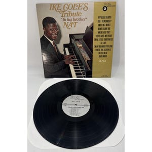 May include: A vintage vinyl record album titled "Ike Cole's Tribute To His Brother Nat." The album cover features a black and white photo of a man playing a piano. The album's tracklist is visible on the right side.