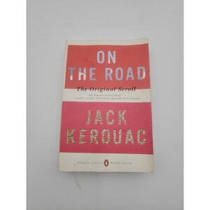 May include: A paperback book titled "ON THE ROAD" by Jack Kerouac. The cover features a red and pink color scheme with white text. The book is a Penguin Classics deluxe edition.