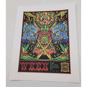 May include: A colorful art print featuring a detailed, symmetrical design with a central figure and crossed rifles. The print includes text that reads "WOOON" and "THE SYLVEE MADISON WI 9/10/23". The artwork is set against a white background.