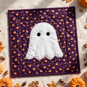 Puede incluir: Un tapiz cuadrado de tela acolchada que presenta un aplique central de fantasma blanco con dos ojos circulares negros, sobre un fondo morado oscuro estampado con pequeñas calabazas naranjas y bordes cosidos.