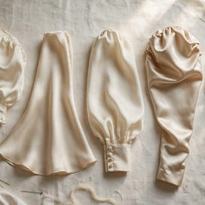May include: A collection of cream-colored satin garment components including a gathered puff sleeve with a ruffled cuff, a flared panel, a long sleeve with a buttoned cuff, a draped sleeve piece, and a small triangular fabric segment.
