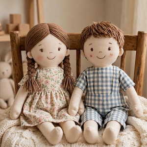 May include: Two fabric dolls sitting side by side. The doll on the left has brown yarn hair in braids and wears a floral print dress. The doll on the right has short brown yarn hair and wears a blue and white gingham overall outfit.