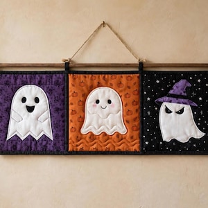 Op de afbeelding: Een gewatteerde wandhanger met drie rechthoekige panelen opgehangen aan een houten roede met touw. De panelen tonen witte spookapplicaties: één op een paarse bloemenachtergrond, één op een oranje pompoenprint en één op een zwarte sterrenachtergrond met een paarse heksenhoed.