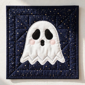 May include: A square quilted wall hanging featuring a white ghost appliqué centred on a dark navy blue fabric background. The navy fabric is patterned with small gold stars. The ghost has black oval eyes, a black mouth, and pink cheeks.
