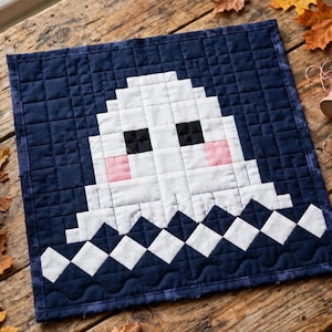 Pode incluir: Uma tapeçaria acolchoada quadrada com um design de fantasma branco pixelado sobre um fundo azul-marinho escuro. O fantasma tem dois olhos quadrados pretos e duas bochechas quadradas cor-de-rosa, com uma fila de formas de diamante brancas na borda inferior.