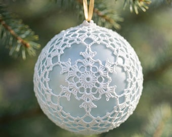 Op de afbeelding: Een bolvormige kerstbal met een lichtblauwe basis bedekt met een wit gehaakt kanten patroon in de vorm van een sneeuwvlok. Het ornament hangt aan een dun gouden lintje aan een dennentak.