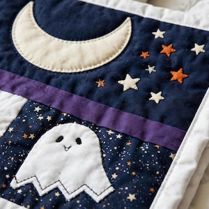 May include: A quilted fabric piece featuring a dark blue background with a cream-colored crescent moon, orange and white stars, and a white ghost appliqué with black embroidered eyes and a small smile, bordered by a white fabric edge.