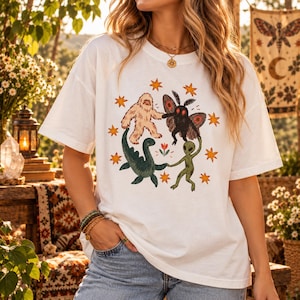May include: A white short-sleeve t-shirt featuring a central graphic print of a beige Bigfoot, a black moth-like creature, a green dinosaur, and a green alien holding hands in a circle, surrounded by orange stars and a small flower.