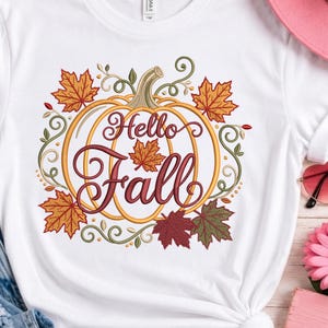 Puede incluir: Una camiseta blanca con un diseño bordado de una calabaza amarilla con contornos dorados. El texto Hello Fall está cosido en letra cursiva de color rojo oscuro en el centro. La calabaza está rodeada de hojas de arce naranjas, rojas y verdes.