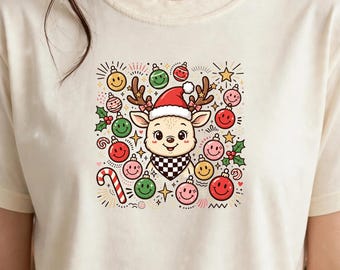 Könnte beinhalten: Ein cremefarbenes T-Shirt mit einer zentralen Grafik eines Cartoon-Rentiers, das eine Weihnachtsmannmütze und ein schwarz-weiß kariertes Halstuch trägt, umgeben von bunten Smiley-Weihnachtskugeln, Sternen, Stechpalmenblättern und einer Zuckerstange.
