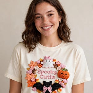 May include: A cream-colored t-shirt featuring a central graphic of a wreath made of crocheted elements including a white ghost with a pink gingham hat, an orange pumpkin, a black bat with a pink bow, candy corn, leaves, and hearts, with the text Spooky Cutie.