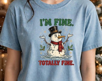 Könnte beinhalten: Ein hellblaues T-Shirt mit einer Grafik eines schmelzenden Schneemanns, der einen schwarzen Zylinder und einen roten Schal trägt. Der Schneemann hat eine Karottennase und Zweigarme. Der Text lautet I'M FINE. und TOTALLY FINE.