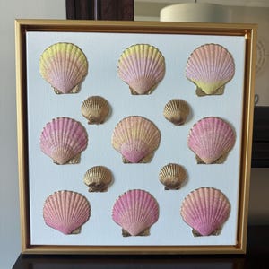 May include: A square wall art piece featuring a white canvas in a gold frame, decorated with twelve seashells arranged in a grid. The shells are painted in gradient shades of pink, yellow, and gold, with some shells featuring a solid metallic gold finish.