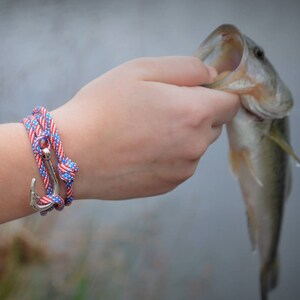 May include: A person wearing a red, white, and blue rope bracelet with an anchor charm. The bracelet is wrapped around the wrist twice. The person is holding a fish with their other hand.