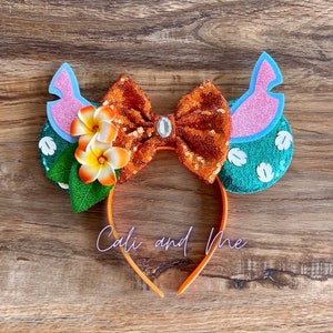 Op de afbeelding: Oranje en groene Minnie Mouse oren met een glimmende oranje strik, witte bloemen en groene bladeren. De oren zijn versierd met een roze en blauw ontwerp. De hoofdband is oranje.
