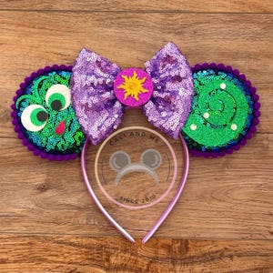 May include: Purple headband with green and purple ears featuring a purple bow with a yellow sun in the center. The ears are decorated with green and purple sequins and have a 3D eye design.