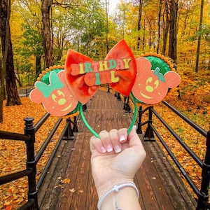 May include: A festive orange and green birthday headband with a large bow and mouse-shaped ears. The bow features the text "BIRTHDAY GIRL" in green letters. The headband is held up against a backdrop of a wooden walkway and autumn foliage.
