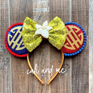 May include: A gold headband with decorative ears featuring a green sequined bow with a white flower accent. The ears are blue and red with gold symbols and pom-pom trim. The text "cali and me" is written on the headband.