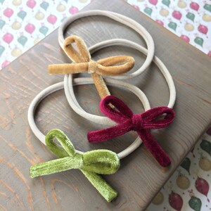 Op de afbeelding: Drie haarbandjes van fluweel met strikken in verschillende kleuren: goud, bordeauxrood en groen. De haarbandjes zijn huidskleur en liggen op een houten oppervlak.