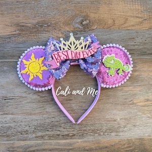 May include: Purple sequined mouse ears headband with a bow that reads "BEST DAY EVER". One ear features a yellow sun, the other a green chameleon. The headband is pink and has the text "Cali and Me".