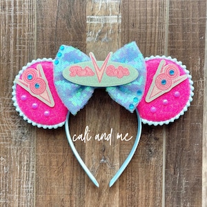May include: A pair of mouse ears with a light blue headband. The ears are hot pink with white trim and a light pink design. The bow is light blue with sequins and the text "Flo's V8 Cafe".