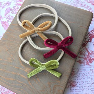 Op de afbeelding: Drie fluwelen haarstrikken in verschillende kleuren: goud, bordeauxrood en groen. De strikken zijn gebonden aan naakte elastische hoofdbanden.