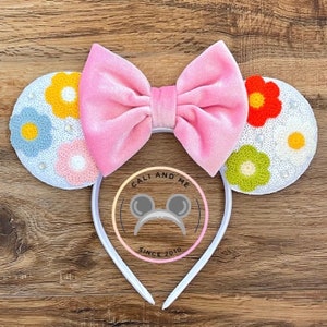 May include: A white headband with mouse ears, a pink bow, and floral embellishments. The ears are white with colorful flower designs and sequins. The bow is a soft pink velvet. The text "CALI AND ME SINCE 2010" is in the center.