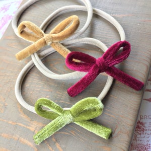 Op de afbeelding: Drie haarbandjes van fluweel met strikken in verschillende kleuren: mosterdgeel, bordeauxrood en olijfgroen. De haarbandjes zijn gemaakt van een rekbaar elastisch materiaal en zijn perfect om haar vast te houden.